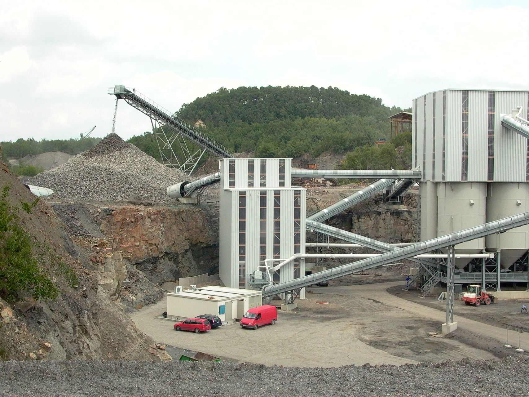 Large gravel and crushed stone dump with several conveyor belts and industrial buildings in a quarry landscape.